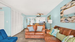 A living room with a brown leather sofa, a blue armchair, and a kitchen in the background. - Gulfstream Condo Unit #401 Heated Pool & Hot Tub | Pickleball Court (Corpus Christi)