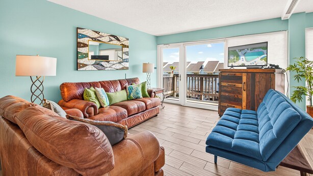 A living room with a brown leather sofa, a blue sofa, a wooden cabinet, and a large window with a view of a pool. - Gulfstream Condo Unit #401 Heated Pool & Hot Tub | Pickleball Court (Corpus Christi)
