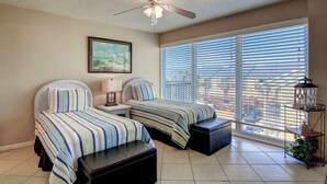 A bedroom with two beds, a ceiling fan, a window with blinds, and a nightstand with a lamp. - Gulfstream Condos Unit 402 | Private Balcony | Pool, Hot Tub & BBQ (Corpus Christi)