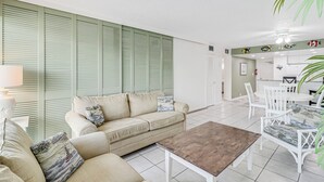 A living room with a beige sofa, a coffee table, and a dining area with white chairs and a table. - Unit 403 Private Balcony/Heated Pool & Hot Tub / BBQ (Corpus Christi)