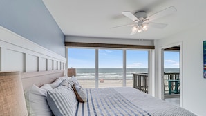 Une chambre avec un grand lit, un ventilateur de plafond et une vue sur la plage, visible à travers de grandes fenêtres.