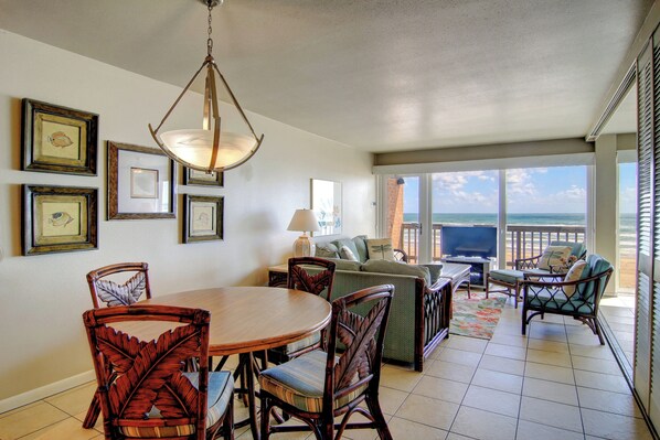 A dining area with a round table and chairs, a sofa, and a TV. There are framed pictures on the wall and a view of the ocean through large windows. - Gulfstream Condos Unit 411 | Gulf Front View | Pool, Hot Tub (Corpus Christi)