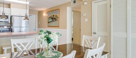 A dining area with a glass-top table, white chairs, and a vase of flowers.