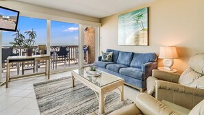 A living room with a blue sofa, beige armchairs, a glass coffee table, and a TV on a stand. - Gulfstream Condos Unit 412 | Gulf- Front View (Corpus Christi)