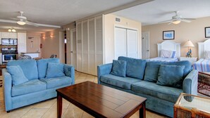A living room with two blue sofas, a wooden coffee table, and a wicker chair. There is a bed with a striped bedspread in the background.
