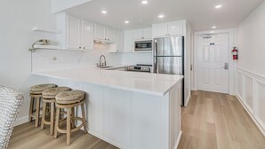 A modern kitchen with white cabinetry, a stainless steel refrigerator, and a central island with stools. - Gulfstream Corner Unit 418 | Wraparound Beachfront Balcony | Pool, Hot Tub (Corpus Christi)