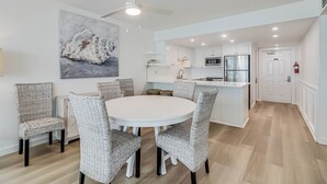 A modern kitchen with white cabinets, a stainless steel refrigerator, and a microwave. There is a dining area with wicker chairs and a white table. - Gulfstream Corner Unit 418 | Wraparound Beachfront Balcony | Pool, Hot Tub (Corpus Christi)