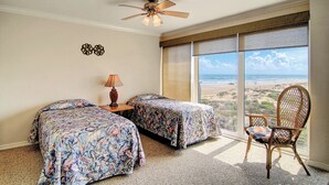 Habitación con dos camas, ventilador de techo, lámpara, una silla y un amplio ventanal con vista al mar.