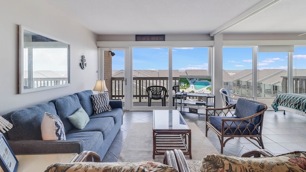 Un salon spacieux avec un canapé bleu, une table basse et des fauteuils en osier. Une grande fenêtre offre une vue sur la plage et les autres bâtiments.