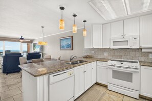 Une cuisine moderne avec des meubles blancs, un plan de travail en granit, un four encastré et un four à micro-ondes. Un coin repas offre une vue sur l’océan.