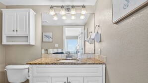 A bathroom with a white vanity, a toilet, a sink with a granite countertop, a mirror, and a ceiling light fixture. - Gulfstream Condos Unit 508 | Gulf Views & Beachfront Balcony | Pool, Hot Tub (Corpus Christi)