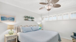 A bedroom with a bed, a ceiling fan, a nightstand with a lamp, a wall-mounted picture, and a window with shutters. - Gulfstream Condos Unit 511 | Gulf-Front Balcony | Pool & Hot Tub (Corpus Christi)