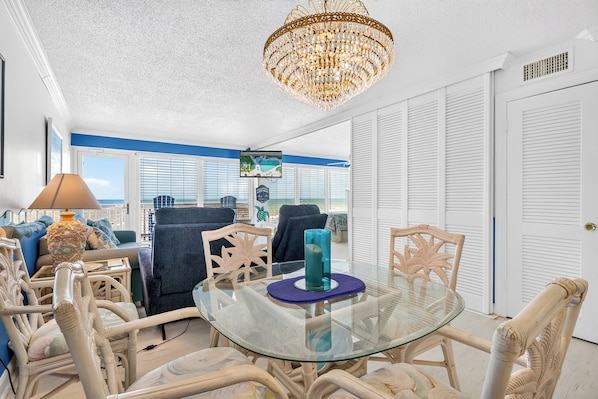 A room with a glass table, wicker chairs, a chandelier, and a TV on the wall.