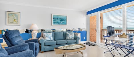 Una sala de estar con un sofá azul, sillones, una mesa de centro y vista al mar a través de amplios ventanales.