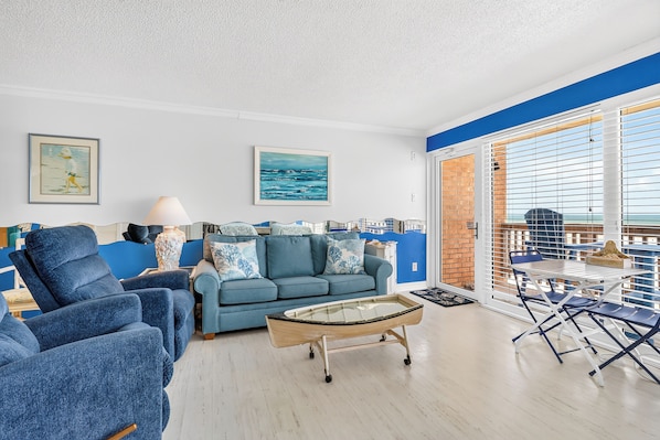 Un salon avec un canapé bleu, des fauteuils, une table basse et une vue sur la mer grâce à de grandes fenêtres.
