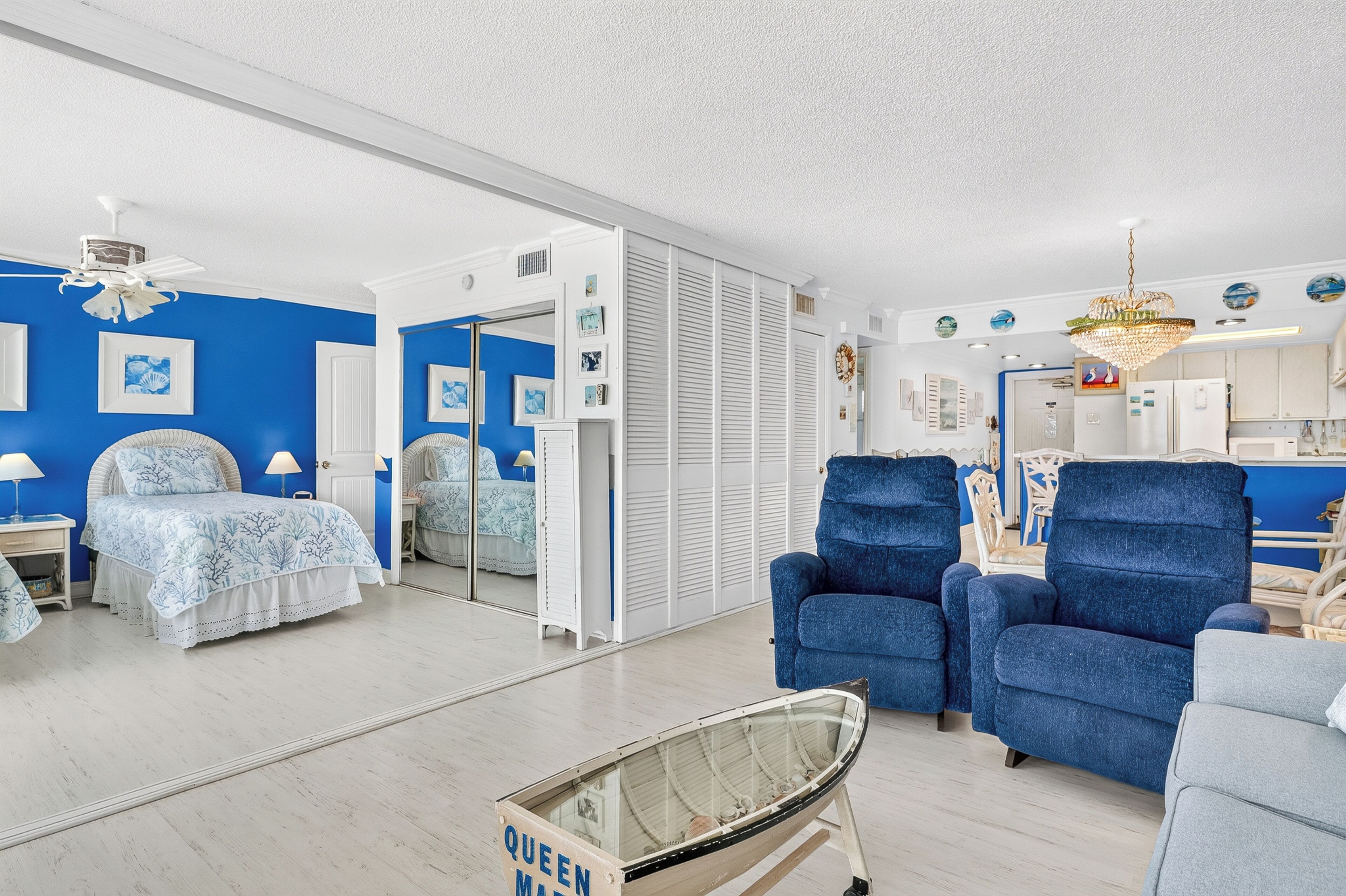 Une pièce comprenant un lit, deux fauteuils bleus, une petite table, un miroir et un ventilateur de plafond.