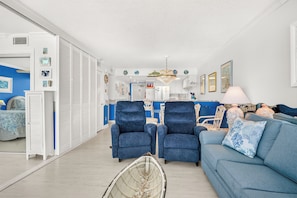 A living room with a blue sofa, two blue armchairs, a dining area with a chandelier, and a kitchen in the background.
