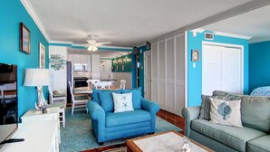 A living room with a blue sofa, a green sofa, and a coffee table. There is a dining table with chairs in the background. - GSC Unit 512 | Pickleball, Pool, Hot Tub & BBQ | Beachfront View (Corpus Christi)