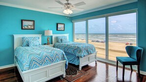 A bedroom with two beds, a ceiling fan, a large window with a beach view, and a chair. - GSC Unit 512 | Pickleball, Pool, Hot Tub & BBQ | Beachfront View (Corpus Christi)