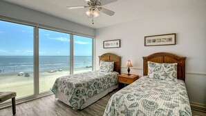Ein Schlafzimmer mit zwei Betten, einem Deckenventilator, Blick auf den Strand und gerahmten Bildern an der Wand.