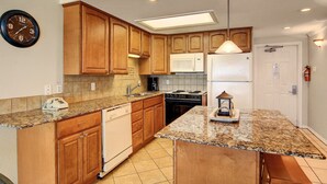 A kitchen with wooden cabinets, granite countertops, a microwave, stove, and a clock on the wall. - Gulfstream Condos Unit 516 | Gulf-Front Balcony | Pool & Hot Tub (Corpus Christi)