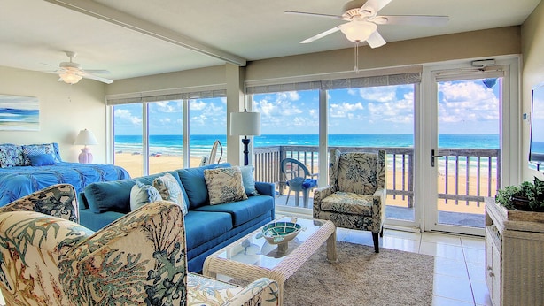 A spacious living room with a large sofa, a coffee table, and a chair. There is a balcony with a view of the beach. - Gulfstream Condos Unit 516 | Gulf-Front Balcony | Pool & Hot Tub (Corpus Christi)