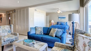 A living room with a blue sofa, floral armchairs, a glass coffee table, and a ceiling fan. - Gulfstream Condos Unit 516 | Gulf-Front Balcony | Pool & Hot Tub (Corpus Christi)