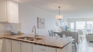 A kitchen with a granite countertop, stainless steel sink, and white cabinetry. There is a dining area with a table and chairs, and a view of buildings through large windows. - Gulfstream Condos Unit 521| Gulf-View Balcony | Heated Pool, Hot Tub (Corpus Christi)