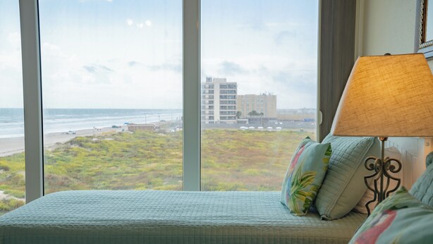 A bedroom with a view of the beach and ocean, a bed with a quilted cover, and a lamp with a beige shade. - Gulfstream Condos Unit 521| Gulf-View Balcony | Heated Pool, Hot Tub (Corpus Christi)