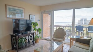 A living room with a TV, sofa, and chairs, a balcony with a view of the beach. - Gulfstream Condos Unit 521| Gulf-View Balcony | Heated Pool, Hot Tub (Corpus Christi)