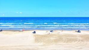 A beach with a clear blue sky, calm sea, and a few people and vehicles on the sand. - Gulfstream Condos Double Unit | 2 Adjoining Beachfront Top-Floor Units (Corpus Christi)