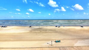 A beach with a clear sky, sandy shore, and a few vehicles. - Gulfstream Condos Unit 616 | Top-Floor | Gulf Front Views (Corpus Christi)