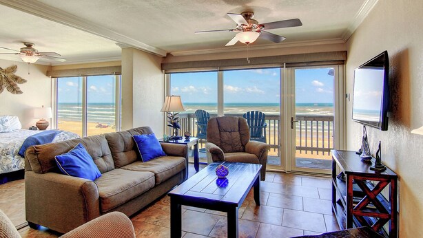 A spacious living room with a large window offering a view of the beach, a ceiling fan, and a flat-screen TV mounted on the wall. - Gulfstream Condos Unit 616 | Top-Floor | Gulf Front Views (Corpus Christi)