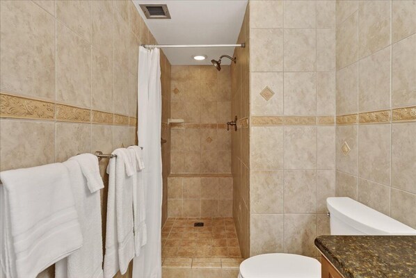 A bathroom with beige tiles, a shower, a toilet, and a sink with a granite countertop.