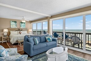 A bedroom with a large window overlooking the beach, a bed, a blue sofa, and a small table.
