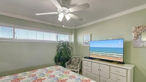 Un dormitorio con cama, cómoda, televisor mostrando una escena de playa, ventilador de techo y ventana con persianas.
