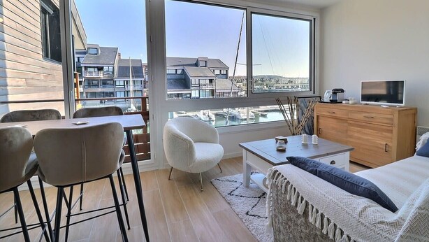 A modern living room with large windows, a sofa, a coffee table, a TV, and a wooden cabinet. - Sublime apartment facing the Port & Planches (Deauville)
