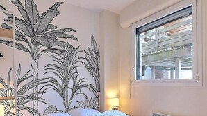 A bedroom with a bed, a window with a view of a wooden structure, a wall with a plant mural, and a bedside table with a lamp. - Sublime apartment facing the Port & Planches (Deauville)