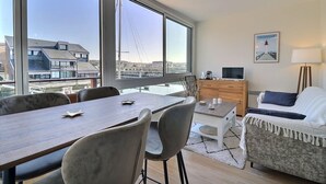A modern living room with a dining area, large windows, a TV, and a cozy seating area. - Sublime apartment facing the Port & Planches (Deauville)