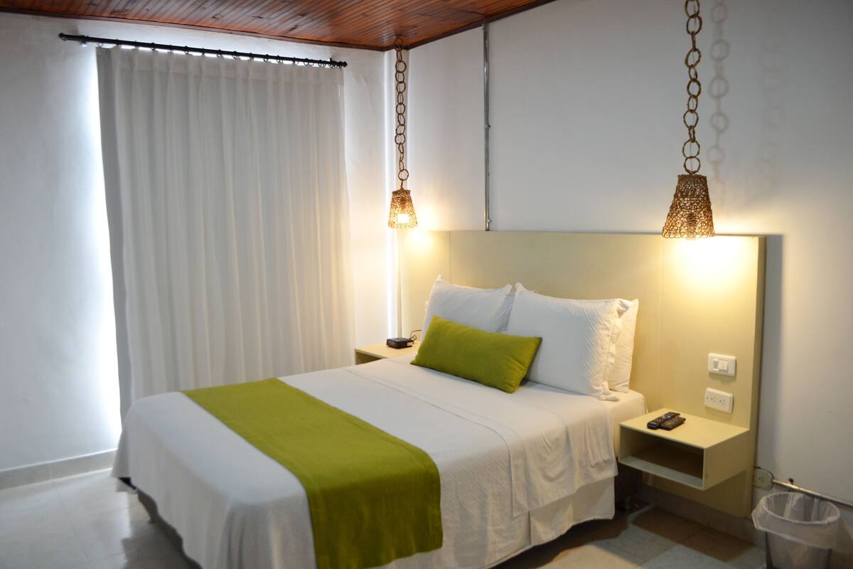 Un lit bien fait, avec du linge de lit blanc et un plaid vert, une table de chevet avec une lampe et une poubelle.