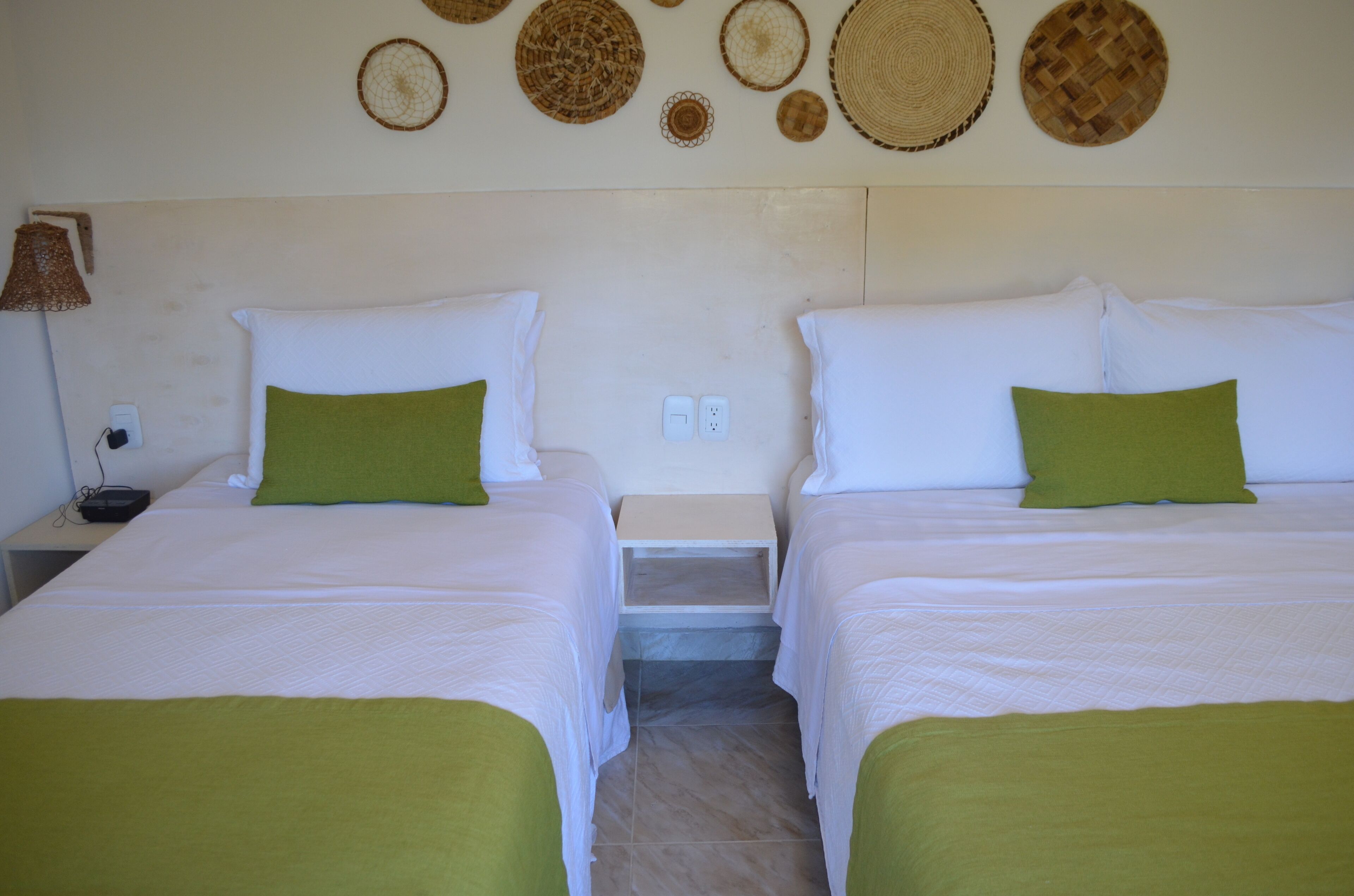 A hotel room with two beds, each with white bedding and green pillows. There are woven wall decorations above the beds and a lamp on the left side.