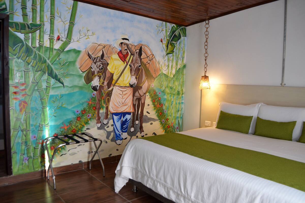 Une chambre d’hôtel ornée d’une fresque représentant un homme et des chevaux, avec un lit doté d’oreillers verts et un luminaire suspendu.