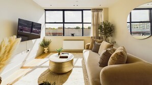 A modern living room with a sofa, a coffee table, and a TV mounted on the wall. There is a large window with a view of the cityscape. - Be London - The St Pancras Residences (London)