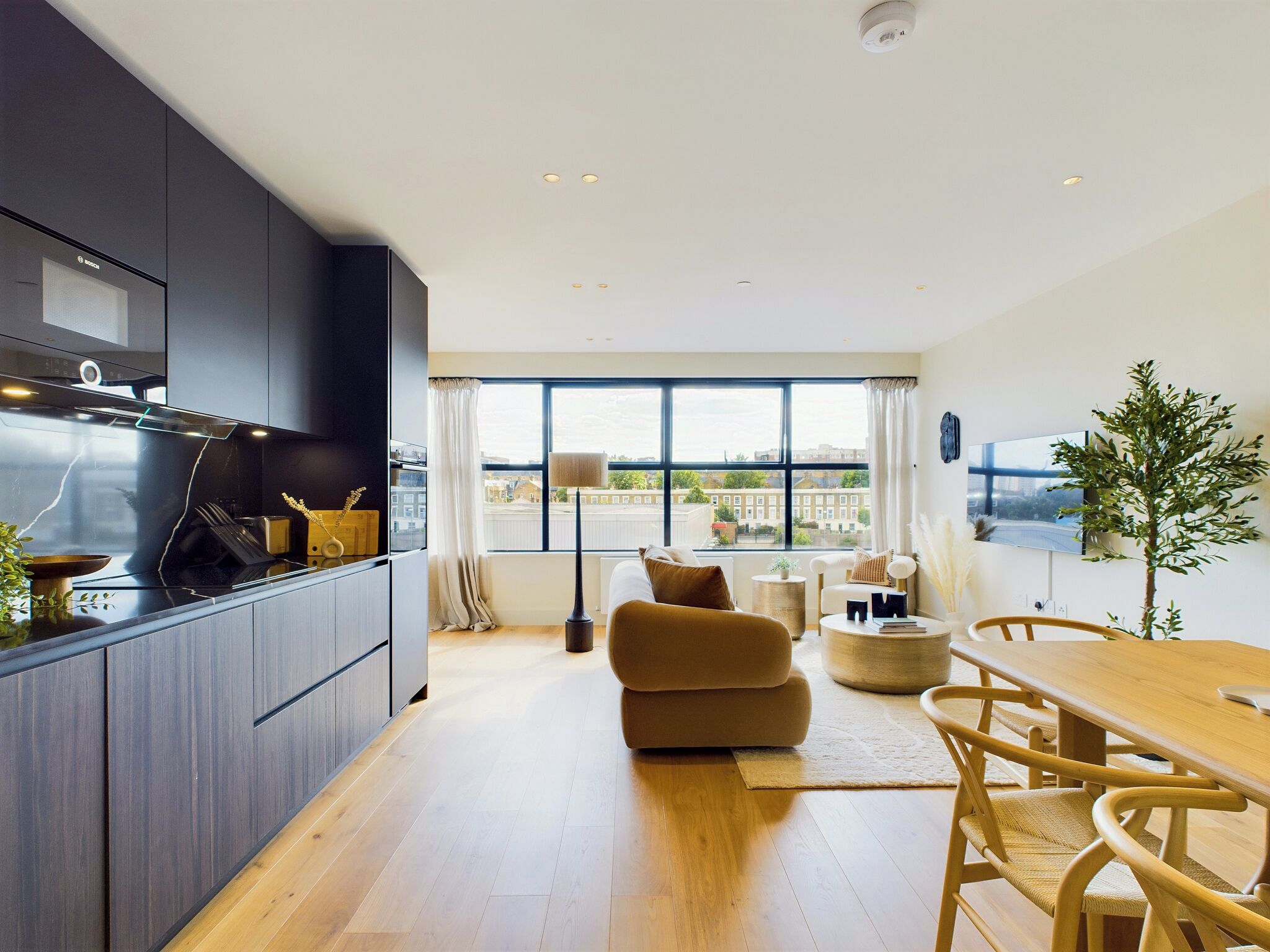 Una cocina moderna con muebles oscuros, una encimera negra y una estufa empotrada. Hay una zona de comedor con una mesa y sillas de madera, un sofá, y un amplio ventanal con vistas a la ciudad.