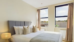 A modern bedroom with a large bed, a bedside lamp, and a view of the city through the windows. - Be London - The St Pancras Residences (London)