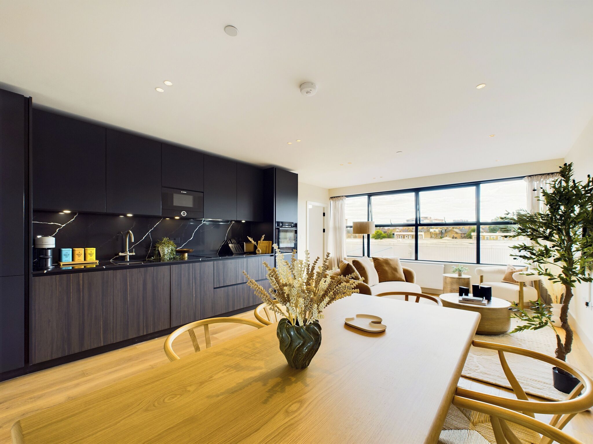 A modern kitchen with dark cabinetry, a wooden dining table, and a large window providing a view of the city.