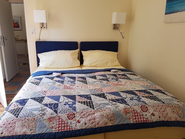 Una cama con colcha de retazos, dos lámparas de noche y una puerta que da a otra habitación.