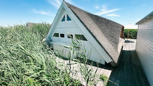 A white building with a sloped roof, surrounded by tall grass and a clear blue sky. - Seehütte Sonnenschilf (Rust)