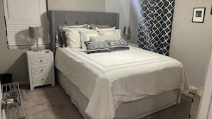 Un dormitorio ordenado con una cama grande, una mesita de noche, una lámpara y una cortina estampada.