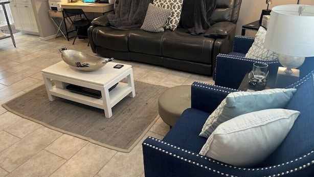 Una sala de estar moderna con un sofá de cuero negro, un sillón azul y una mesa de centro blanca. Se observa un escritorio con una computadora portátil y un monitor de computadora al fondo.